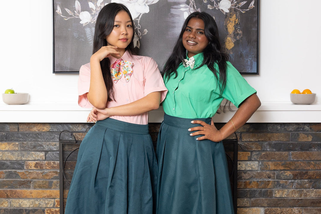 Two women wearing matching outfits with green tops and blue skirts in a kitchen setting.