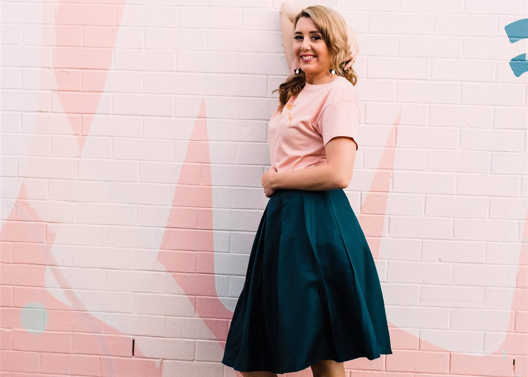 Woman in a pink shirt and teal skirt standing against a pink brick wall.