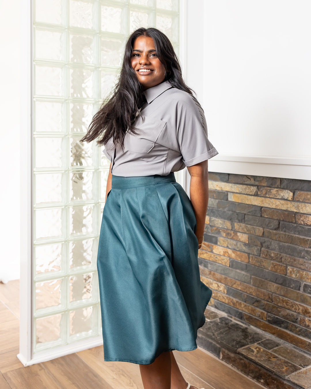 Woman wearing a gray shirt and teal skirt standing in front of a brick wall.