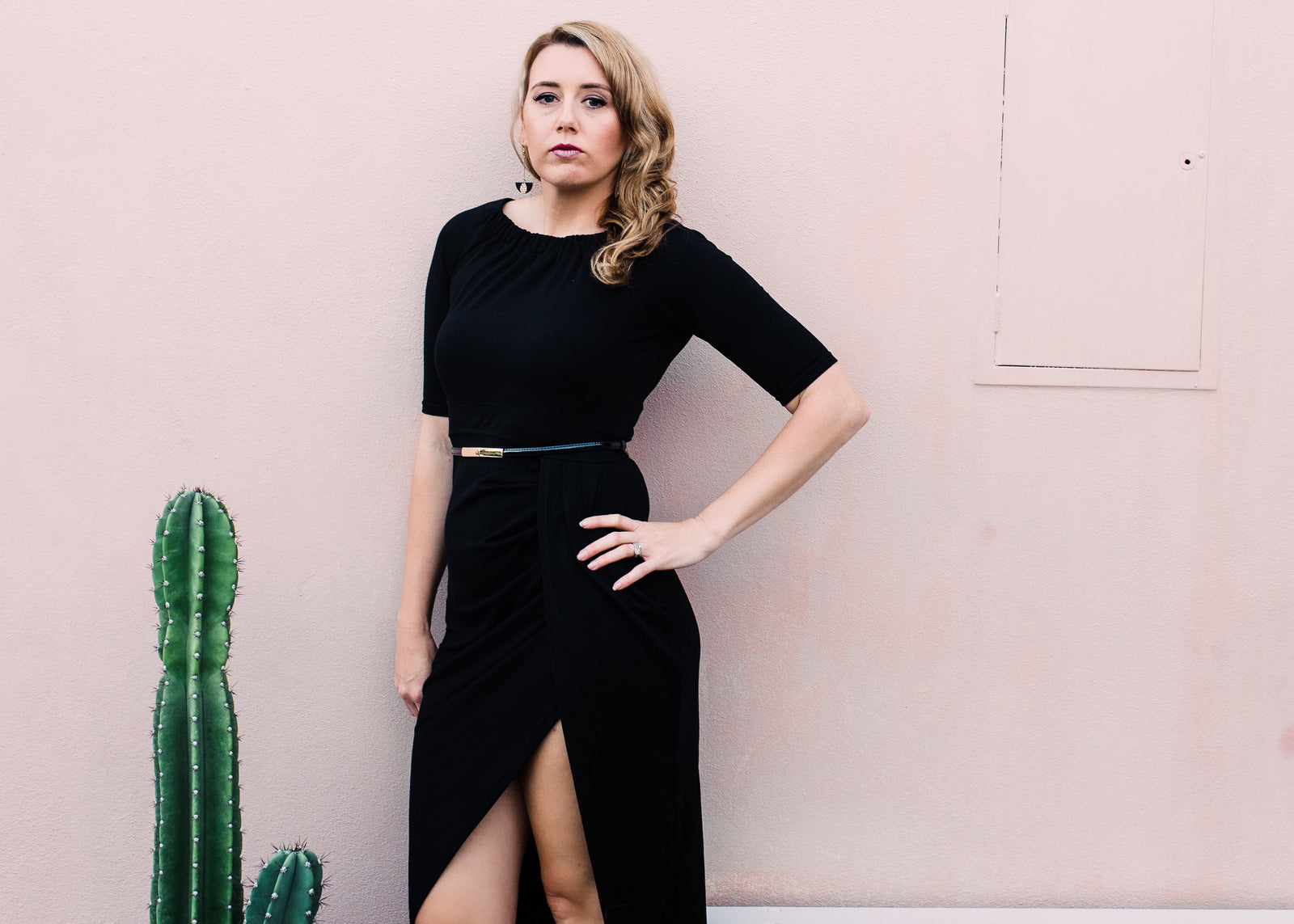 Woman in a black dress standing next to a cactus plant against a pink wall.
