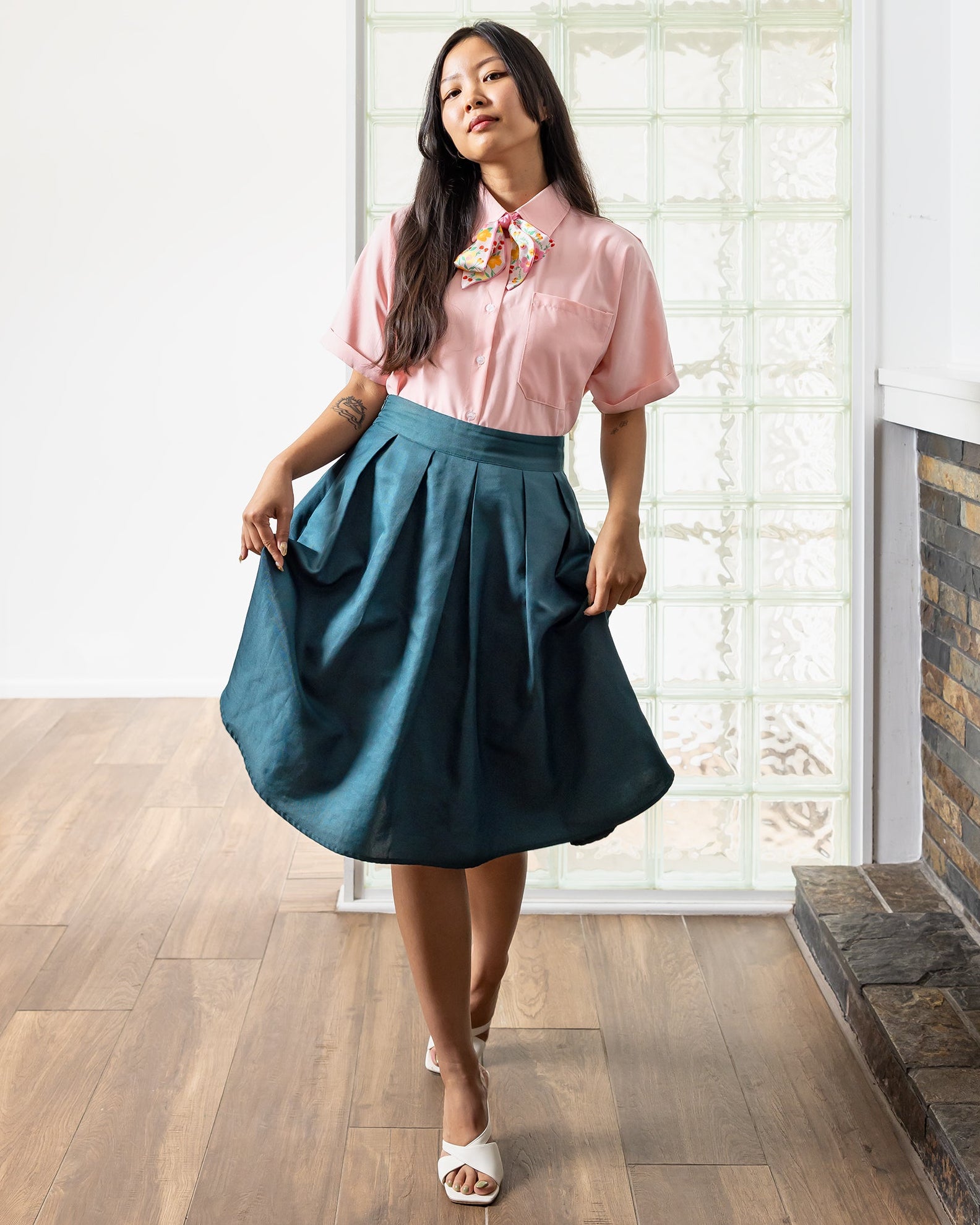 Woman in a pink shirt and teal skirt standing in a room with wooden flooring and a stone wall.